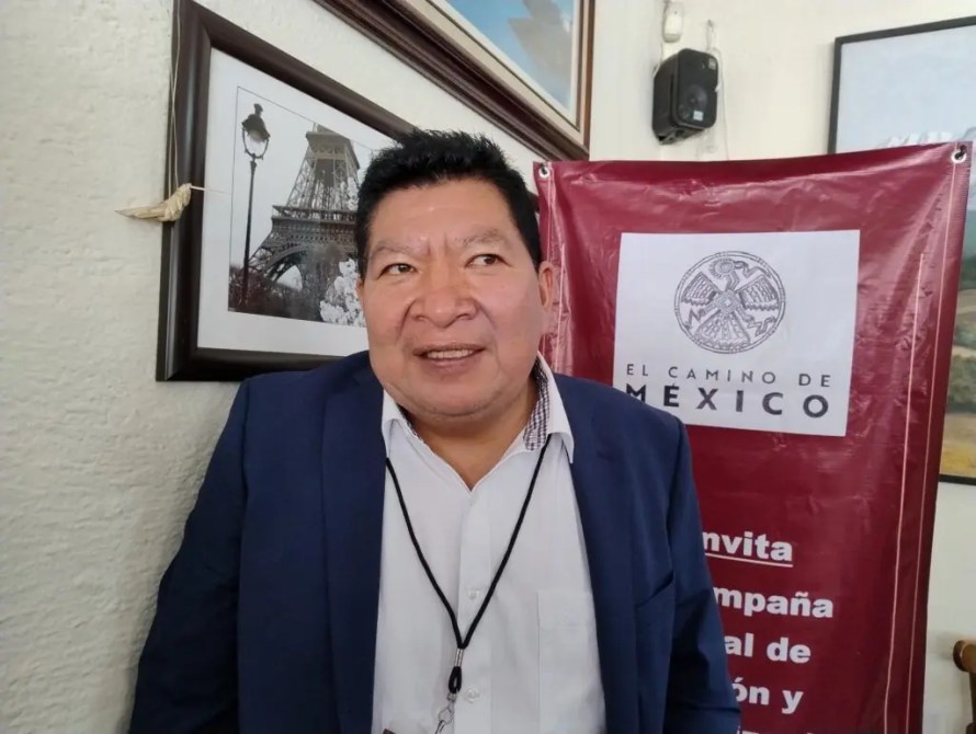 A man in a dark blazer and patterned shirt stands in front of a red banner that reads 'EL CAMINO DE MEXICO.' Behind him are framed photographs, including one of the Eiffel Tower.
