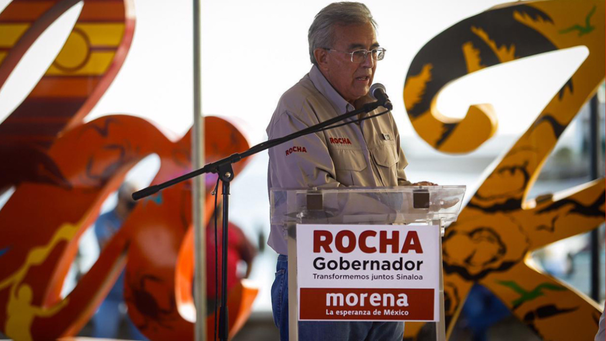 Un hombre, vestido con una camisa clara que tiene la palabra 'ROCHA', habla en un podio con un micrófono. Detrás de él, hay letras decorativas en colores cálidos. El podio tiene un cartel que dice 'ROCHA Gobernador'.