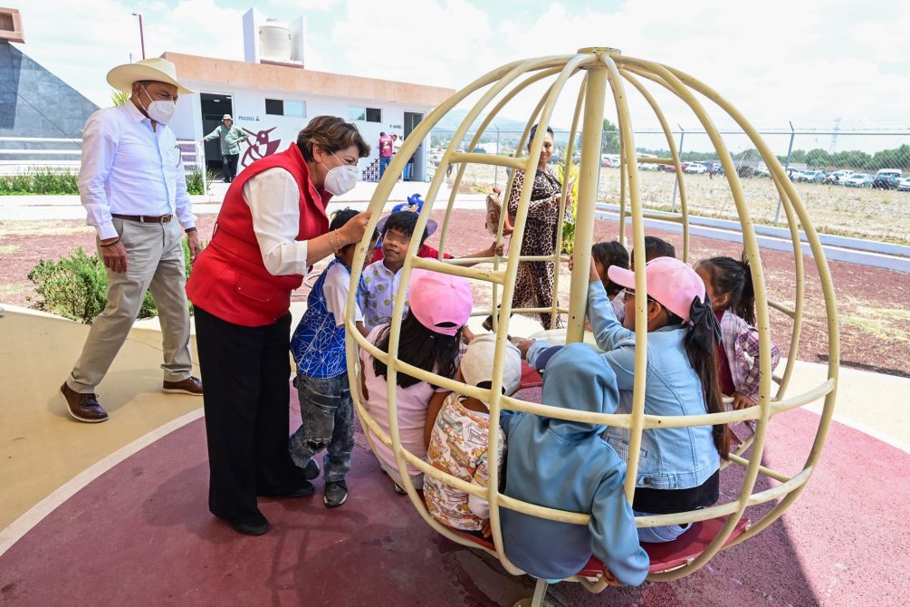 A group of children playing in a round, cage-like structure while an adult engages with them, set in an outdoor playground.