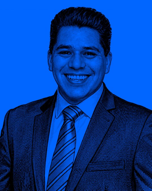 Retrato de un hombre sonriente con traje oscuro y corbata, sobre un fondo azul.