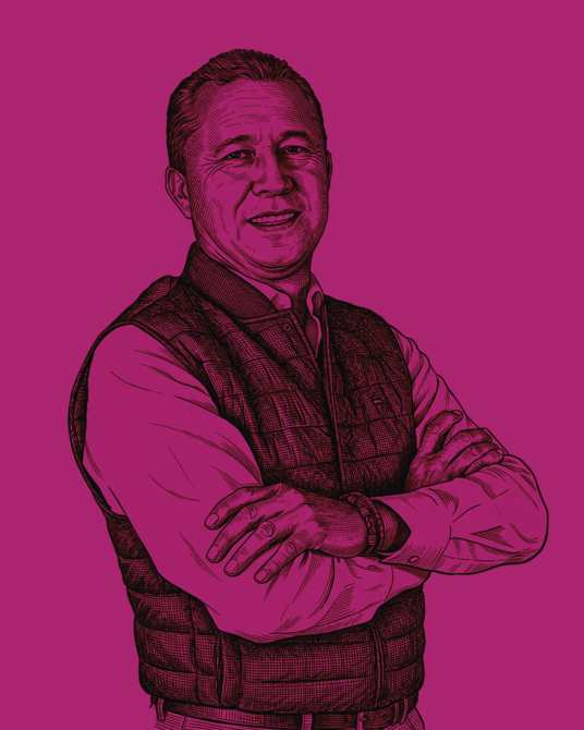 Retrato de un hombre sonriendo con brazos cruzados, vestido con un chaleco sobre una camisa blanca, con un fondo de color magenta.