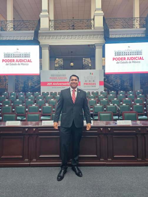 Un hombre de pie en un salón con sillas verdes, frente a pantallas que muestran el texto 'Poder Judicial del Estado de México' y '201 Aniversario'.