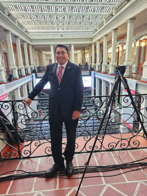 Hombre de pie en un balcón con barandilla, vistiendo un traje negro y corbata a rayas rojas y blancas, con una cámara en un trípode al lado, en un edificio con un diseño arquitectónico elegante.