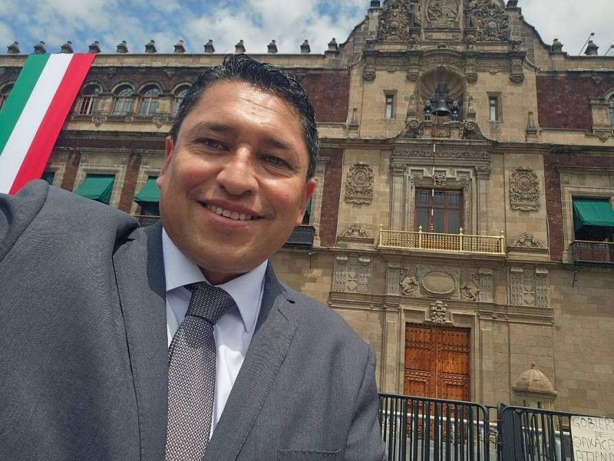 Autorretrato de un hombre sonriente en frente de un edificio histórico, con una bandera mexicana al fondo.