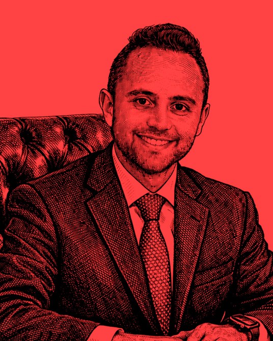 Retrato de un hombre vestido formalmente, sonriendo, sentado en una silla de cuero con un fondo rojo.