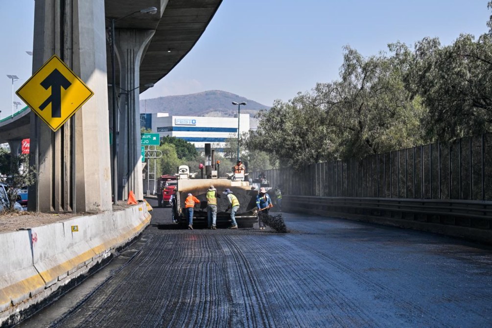 Trabajadores realizando reparación de carretera bajo un puente, con señal de desvío en la esquina.