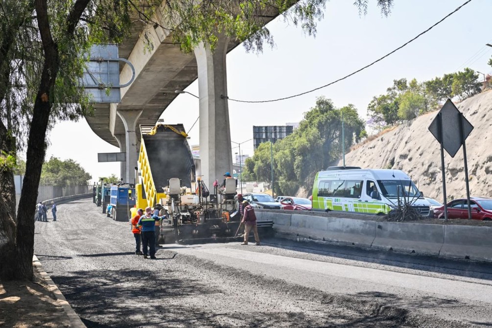 Trabajadores realizando reparaciones en una carretera bajo un puente, con maquinaria pesada y vehículos en la vía.