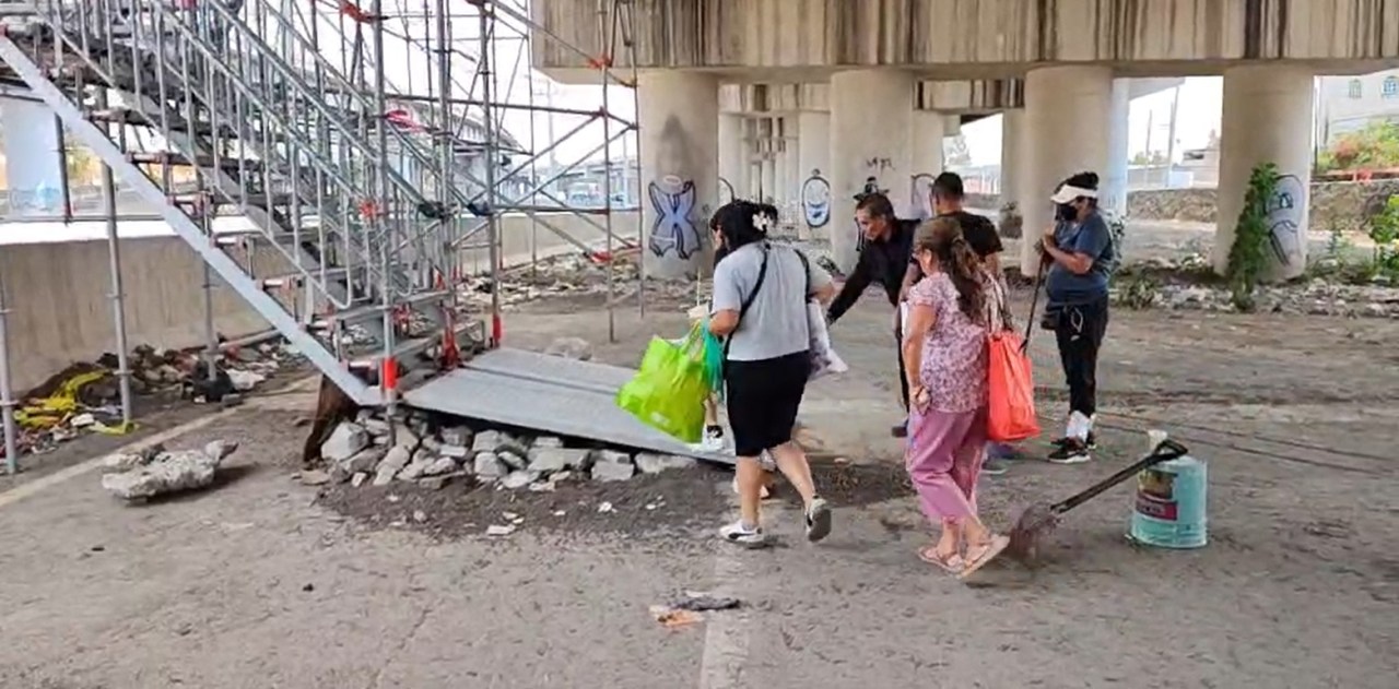 Grupo de personas caminando bajo una estructura de andamiaje cerca de unas escaleras mientras cargan bolsas de compras.