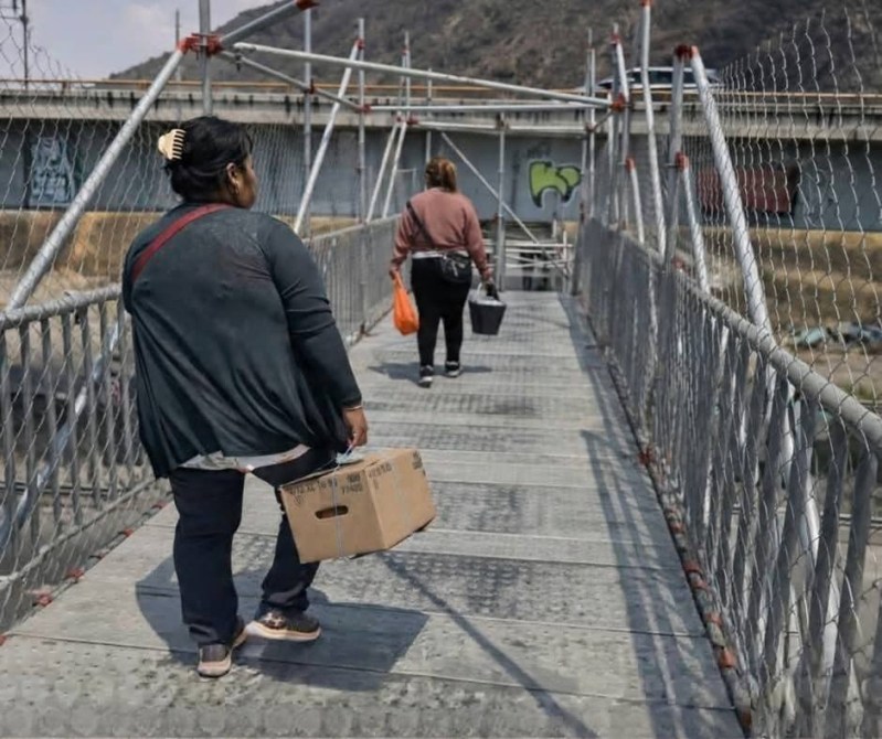 Dos mujeres cruzando un puente colgante, una llevando una caja y la otra una bolsa de plástico.