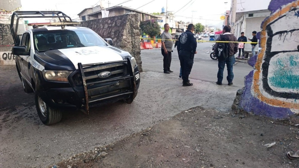 Un vehículo policial estacionado en una calle. Varias personas se congregan en el área, algunas conversando, mientras la escena está delimitada con cinta de precaución y hay graffiti en una pared cercana.