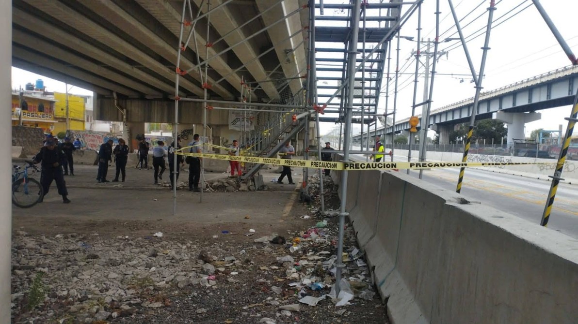 Escena de un sitio de construcción bajo un puente, con trabajo en andamios y presencia policial, rodeado de cintas de precaución y basura en el suelo.