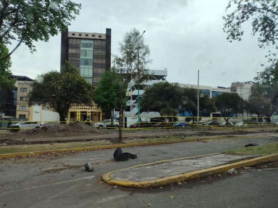 Vista de una calle en construcción con edificios al fondo y árboles a los lados.