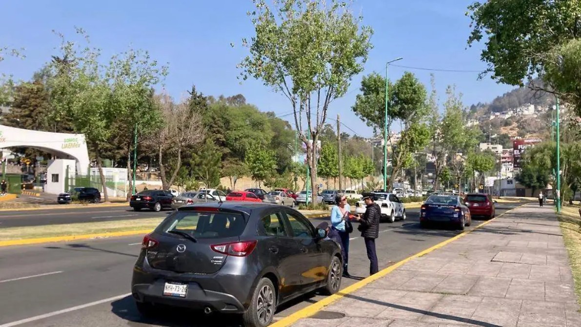 Una vista de una calle con árboles y vehículos estacionados, dos personas conversando en la acera, y un vehículo policial en la parte trasera.
