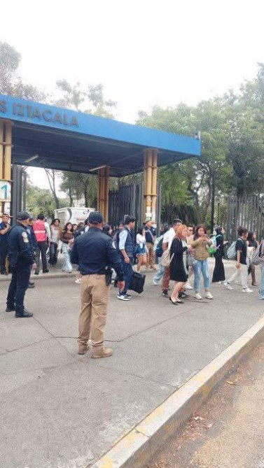A crowd of people gathered at the entrance of a campus, with visible security personnel in uniform. Trees are in the background, and the sky is overcast.