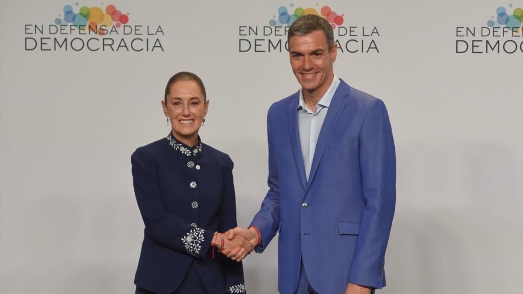 Two individuals shaking hands in front of a backdrop with the text 'En Defensa de la Democracia'. One is wearing a dark suit with floral accents, and the other is in a light blue suit.