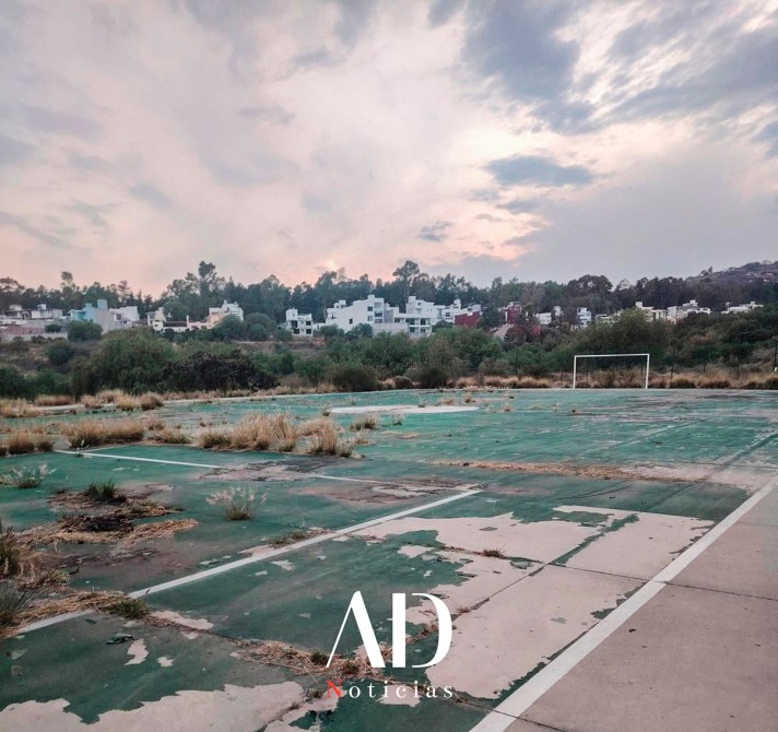 Vista de una cancha de fútbol en desuso con césped seco y descuidado, rodeada de vegetación y edificios al fondo, bajo un cielo nublado.