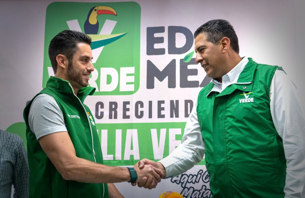 Dos hombres en chaquetas verdes estrechan manos durante un evento relacionado con el partido Verde en México, con un fondo que incluye el logo del partido.