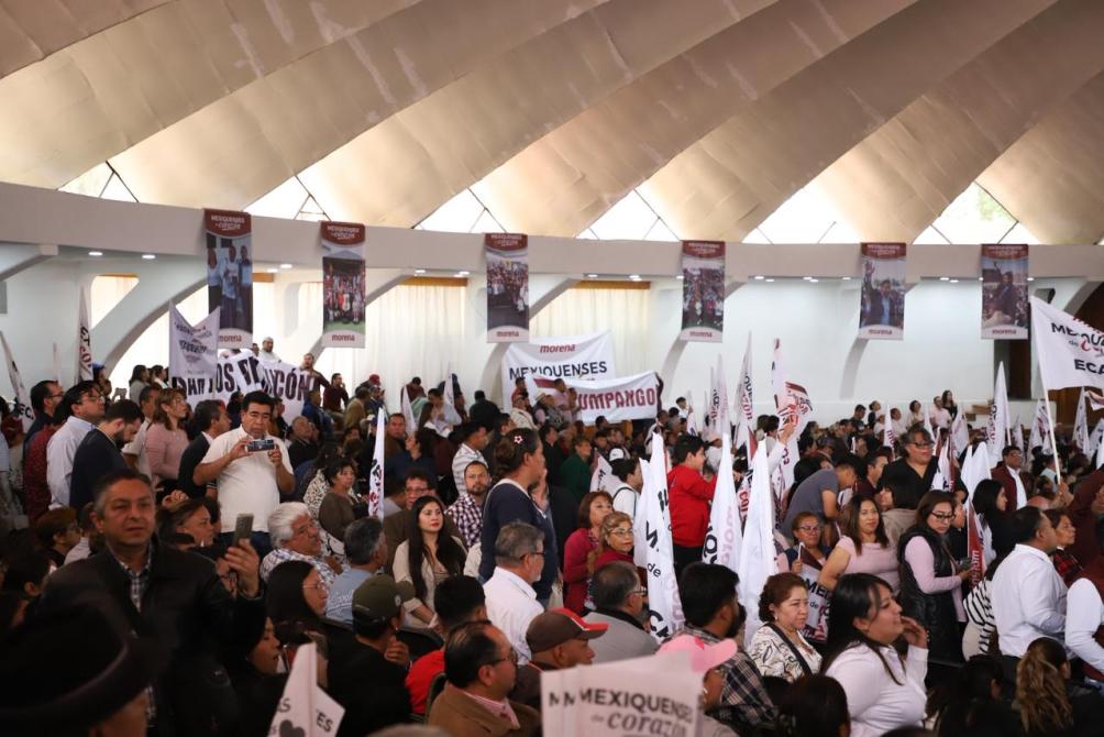 Una gran multitud de personas reunidas en un evento, sostenido banderas y carteles, dentro de un recinto con techo arqueado.