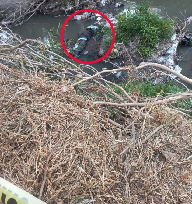 Cuerpo parcialmente sumergido en un canal, rodeado de vegetación y desechos.