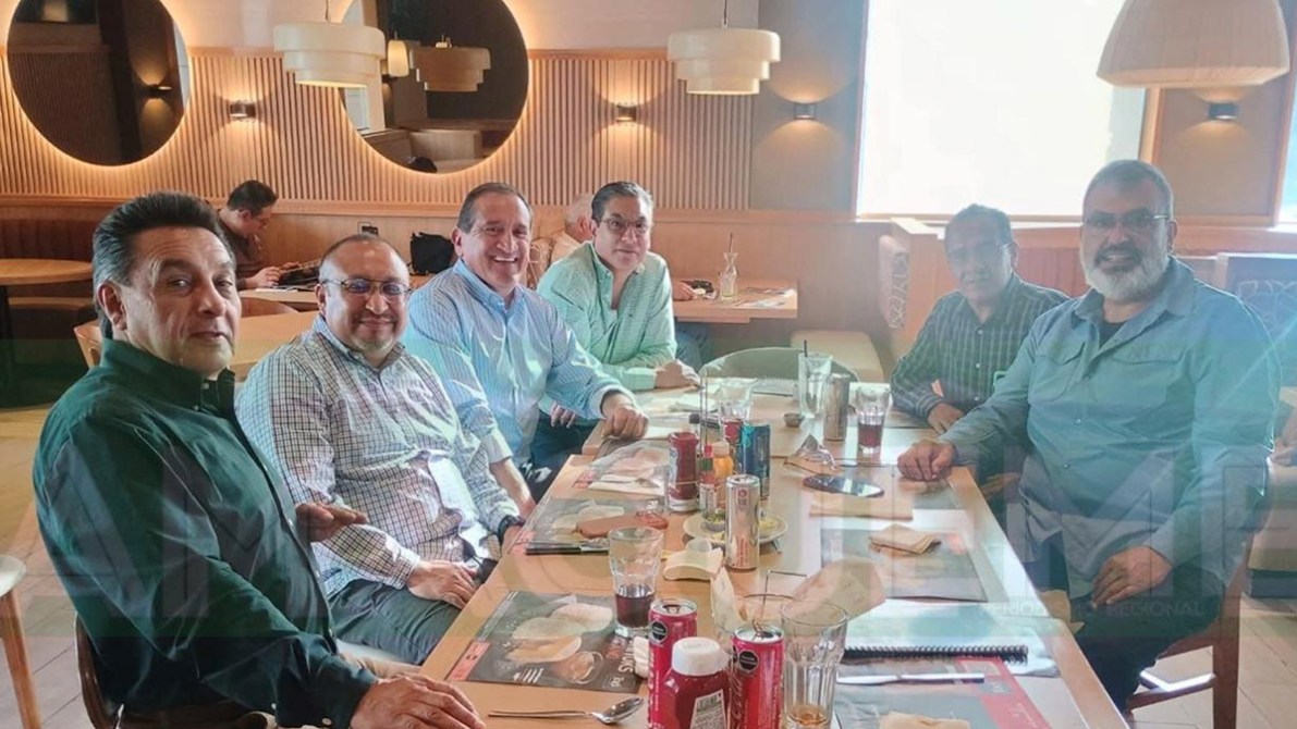 Grupo de seis hombres sentados alrededor de una mesa en un restaurante, sonriendo y conversando. Hay bebidas y comida en la mesa.