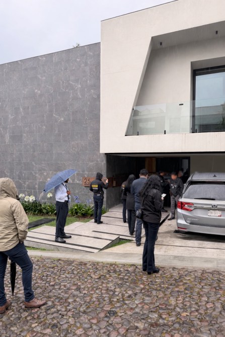 Grupo de personas frente a una residencia moderna, algunos con paraguas, otros entrando. Hay un vehículo estacionado en la entrada.