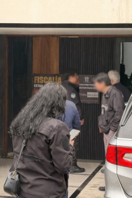 Una mujer con cabello rizado sostiene un teléfono móvil frente a un edificio de la fiscalía, donde varias personas están de pie, algunas de ellas con chaquetas oscuras.