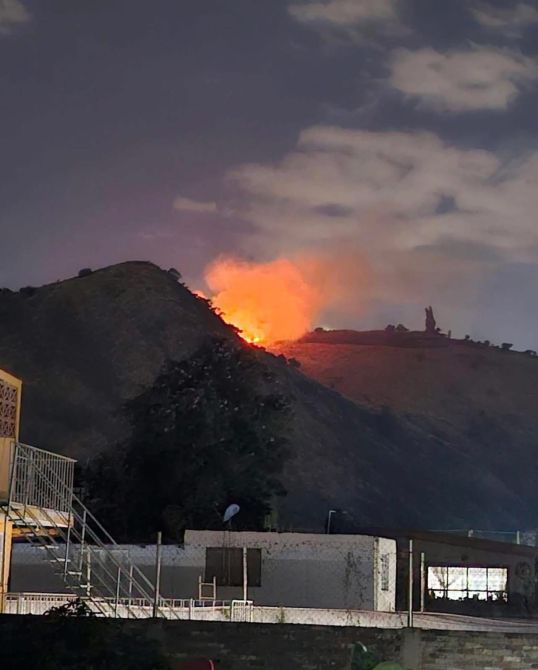 Incendio visible en la cima de una colina durante la noche, con llamas brillantes y un cielo nublado.