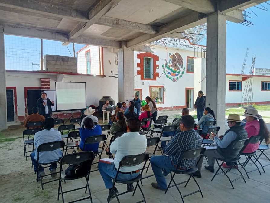Reunión comunitaria en un espacio semiabierto, con un presentador hablando frente a una audiencia sentada en sillas. Se observa un proyector y una pantalla en el fondo.