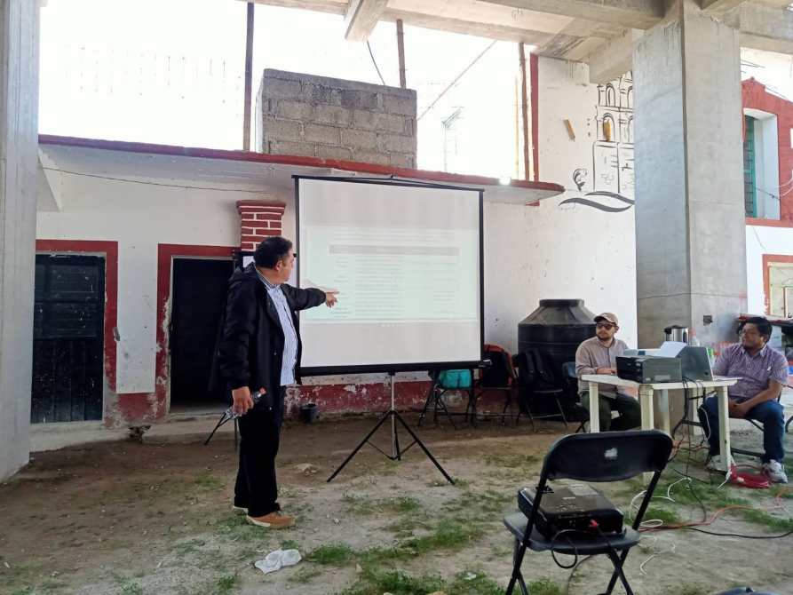 Un presentador señala una diapositiva en una pantalla durante una reunión en un espacio interior con paredes de ladrillo expuesto. Dos personas sentadas a una mesa observan la presentación.