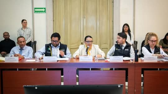 Reunión de varias personas sentadas en una mesa, discutiendo temas importantes. Al fondo se pueden ver otras personas observando.