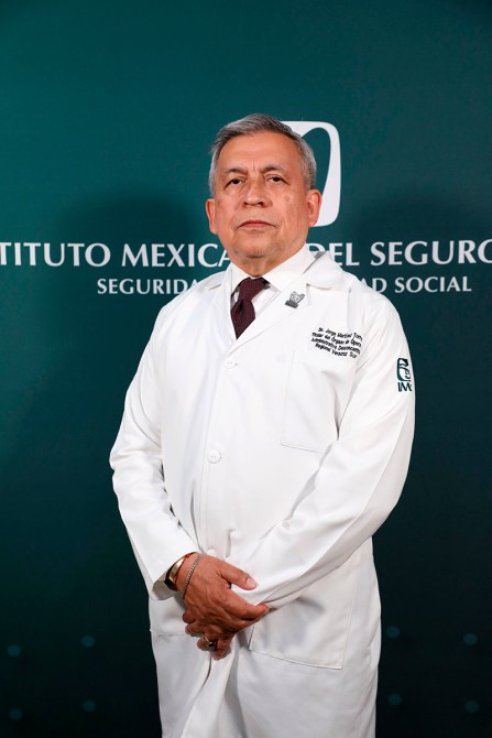Médico de pie, vestido con bata blanca, ante un fondo con el logotipo del Instituto Mexicano del Seguro Social.