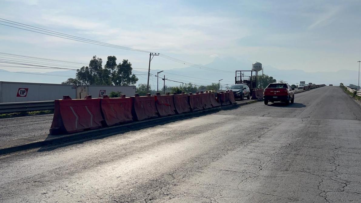 Vista de una carretera con barreras de concreto, vehículos en movimiento y un paisaje montañoso al fondo.