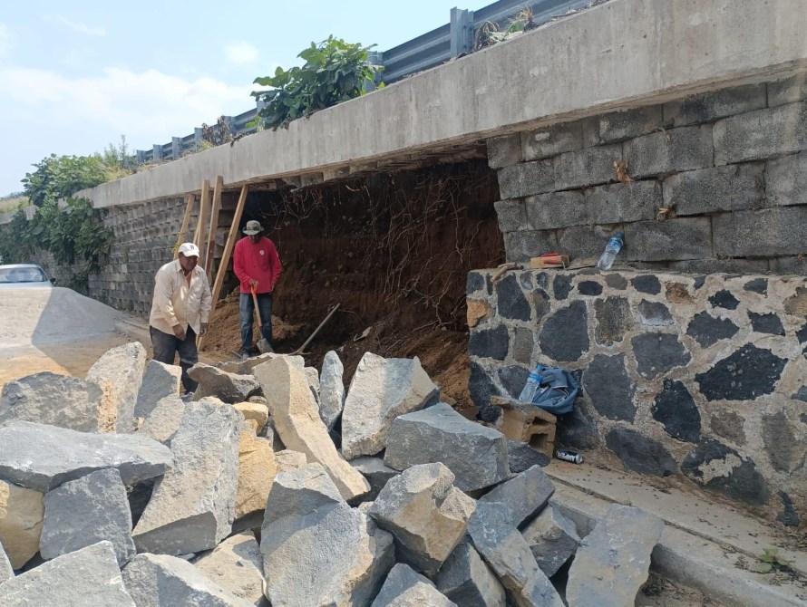 Dos trabajadores excavando junto a un muro de contención, con piedras grandes apiladas en el suelo.