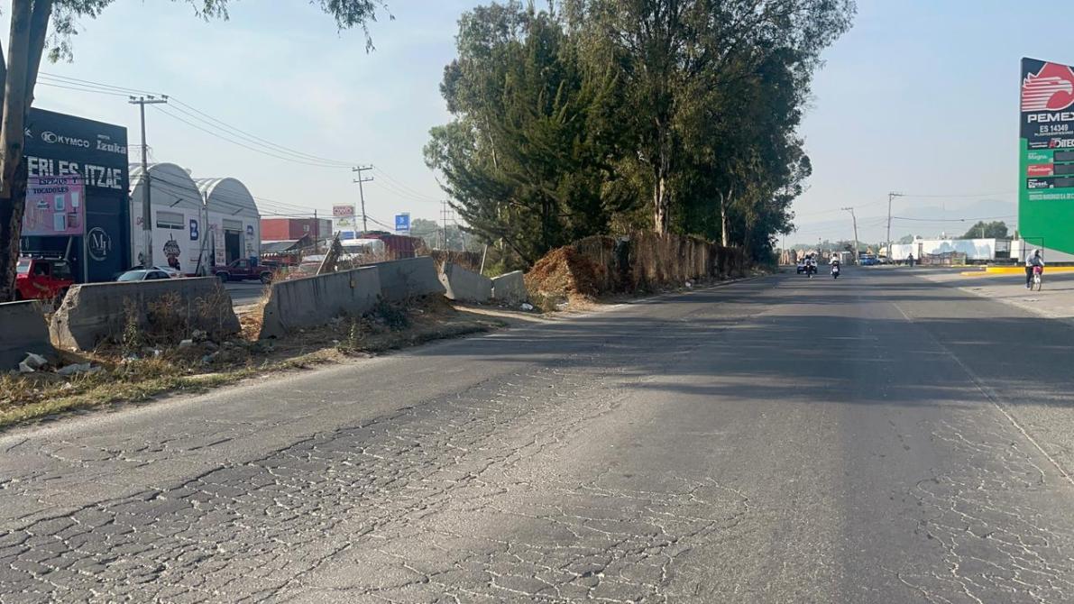 Vista de una carretera con bacheo visible, donde se pueden ver vehículos estacionados y otros en movimiento, además de un letrero comercial a la derecha.