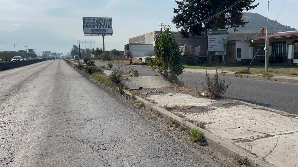 Vista de una carretera con pavimento agrietado, con vehículos en la vía y vegetación crecida en el arcen. Se observa un cartel publicitario en el fondo.