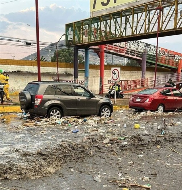 Vehículos atrapados en una calle inundada con basura y escombros, bajo un puente peatonal en un entorno urbano.