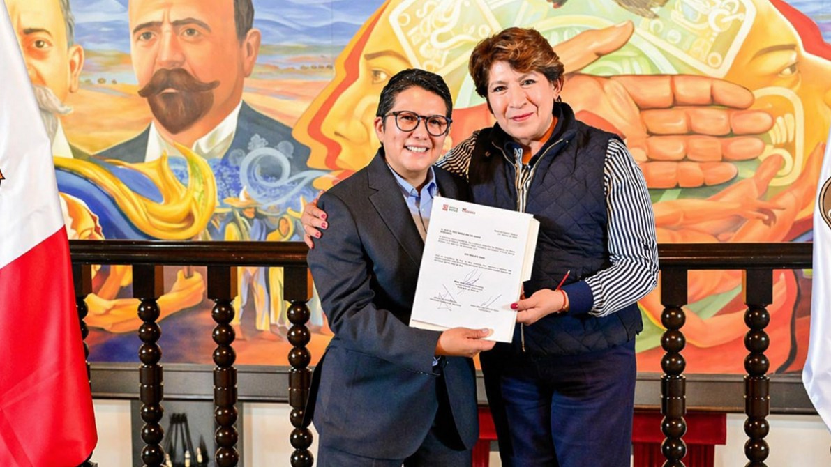 Una mujer y un hombre se están dando la mano mientras sostienen un documento, con un mural colorido de fondo. La mujer viste un chaleco oscuro y blusa a rayas, mientras que el hombre lleva una camisa y chaqueta. Banderas de México están visibles en la imagen.