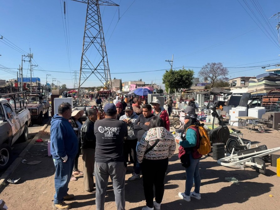 Grupo de personas reunidas en un mercado al aire libre, con varios vehículos y objetos a su alrededor, bajo un cielo claro.