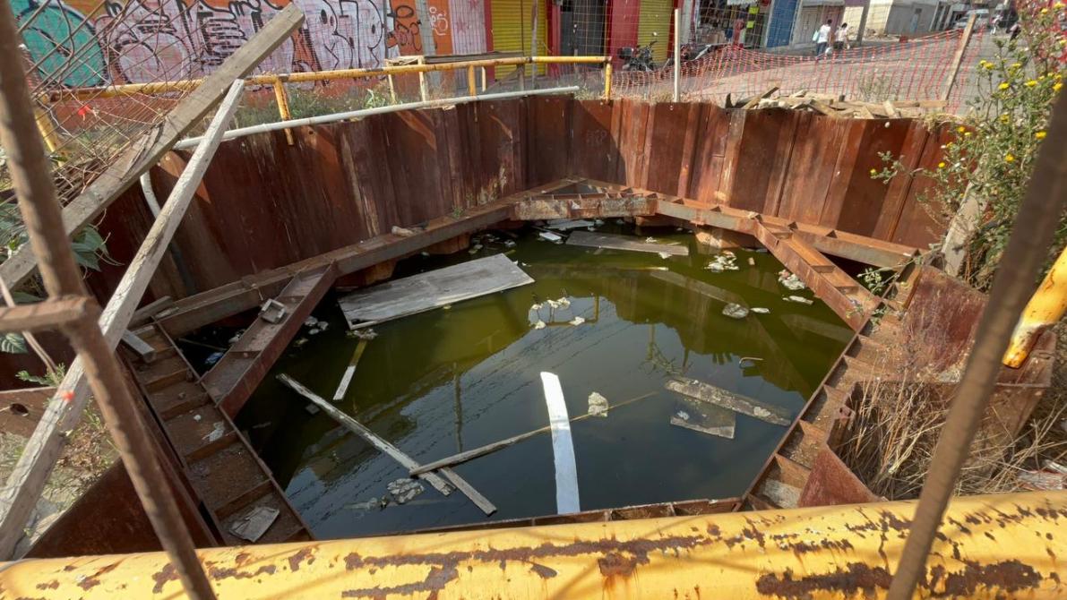 Un agujero cuadrado con agua sucia y desechos en su interior, rodeado de paredes de metal oxidado y vegetación en los bordes.