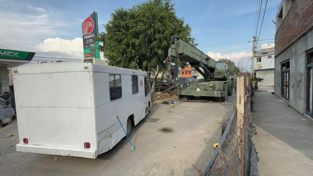Camión blanco estacionado junto a una grúa militar en una calle urbana, con un árbol al fondo y un cartel de PEMEX visible.