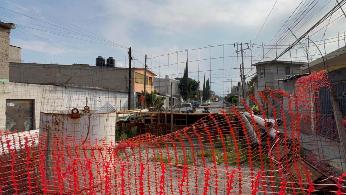 Vista de una calle con una zanja cerrada por una malla de seguridad naranja y un tanque de almacenamiento.