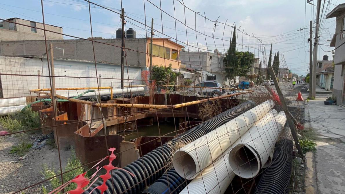 Vista de una calle en construcción con tuberías y una cerca de alambre. Al fondo, se pueden ver casas y un camino pavimentado.