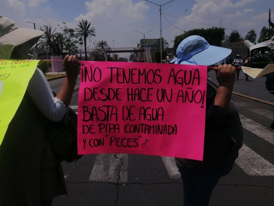 Persona sosteniendo un cartel rosa que dice '¡NO TENEMOS AGUA DESDE HACE UN AÑO! BASTA DE AGUA DE PIPA CONTAMINADA Y CON PECES' en una manifestación.