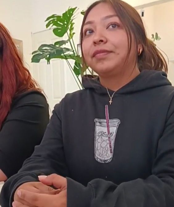 Mujer joven sentada en una mesa, luciendo un hoodie negro con un diseño de vaso. Muestra una expresión pensativa mientras observa. Al fondo, se pueden ver plantas decorativas.