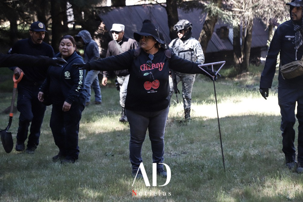 Grupo de personas en un bosque, algunos llevan herramientas como palas. Una mujer destaca en el centro con un sombrero y una camiseta que dice 'Cherry', sosteniendo un dispositivo en forma de triángulo.