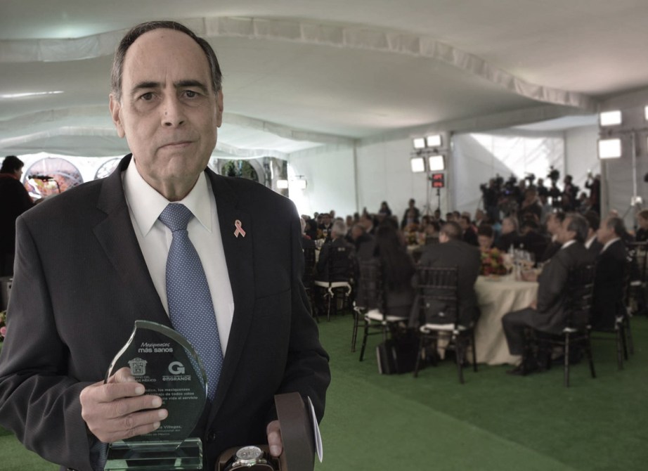 Hombre en traje sosteniendo un trofeo y un reloj, en un evento formal con mesas y asistentes al fondo.