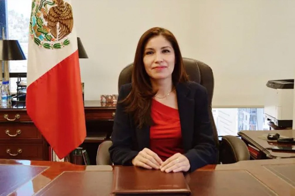 Mujer sentada en un escritorio con una blusa roja y un saco negro, sonriendo. Detrás de ella hay una bandera de México y un ambiente de oficina.