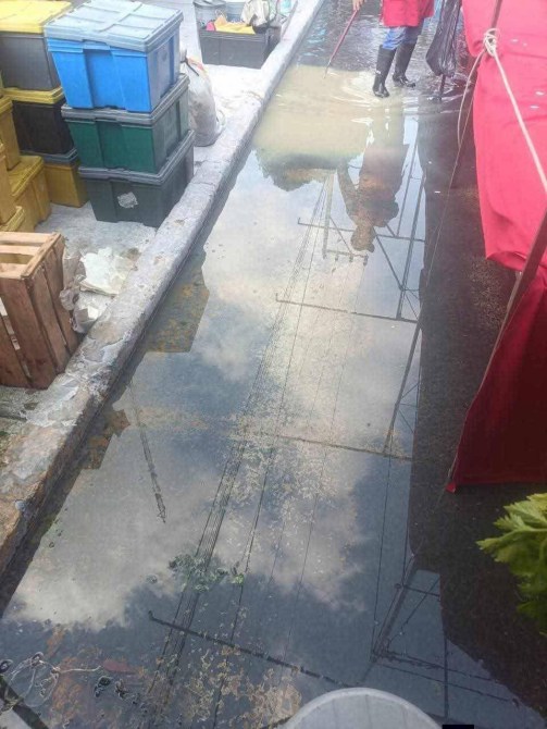 Vista de un canal de agua estancada en un mercado, con reflejos del cielo y cables eléctricos visibles. Hay cajas y algunos desechos alrededor.