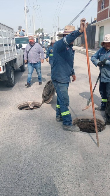 Trabajadores realizando mantenimiento en alcantarillas en una calle, con un vehículo de trabajo y varias personas observando.