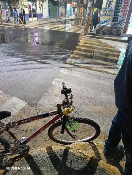 Bicicleta estacionada al borde de una calle húmeda, con reflejos visibles en el pavimento y una persona de pie cerca.